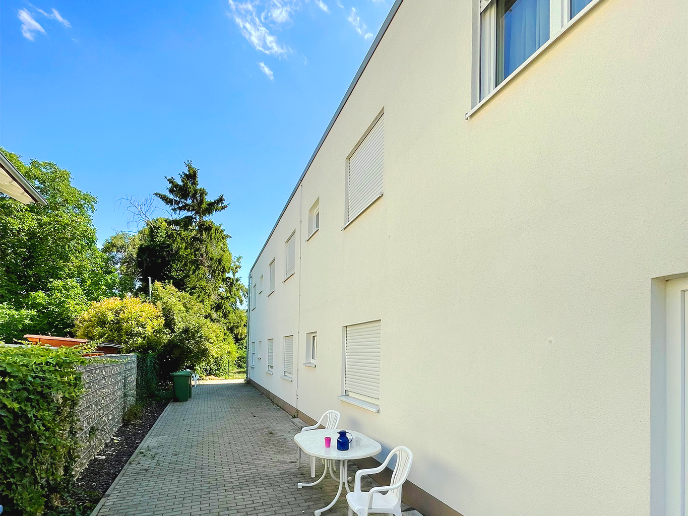 Frisch renovierte Fassade eines Wohngebäudes mit heller Putzoberfläche, gepflastertem Weg und einfacher Sitzgelegenheit