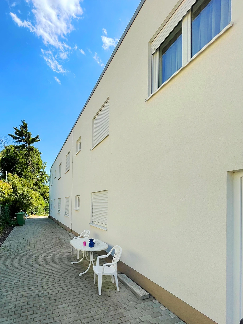 Frisch renovierte Fassade eines Wohngebäudes mit heller Putzoberfläche, gepflastertem Weg und einfacher Sitzgelegenheit